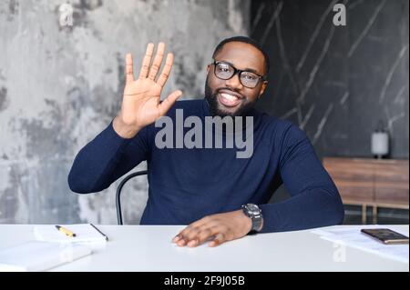 Joyeux homme noir multiracial souriant assis au bureau, regardant l'appareil photo, en agitant la main en disant bonjour pendant un appel vidéo. Webcam portrait d'un coach d'affaires, chef de la direction, consultant financier ou professeur d'université Banque D'Images