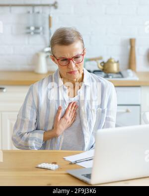 Triste malade femme d'âge moyen souffrant de douleur dans la poitrine ou toux ayant consultation en ligne avec le médecin sur ordinateur portable pendant l'épidémie de Covid 19, assis dans la cuisine à la maison. Concept de santé et de médecine Banque D'Images