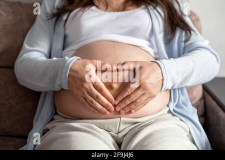 La femme enceinte avec le grand ventre avancé grossesse se reposant sur un canapé à la maison faisant les mains du cœur Banque D'Images