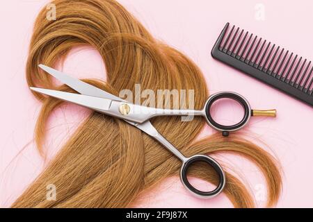 Ciseaux, un peigne et une serrure de cheveux de femme blond bouclé sur un fond rose, vue du dessus. Banque D'Images