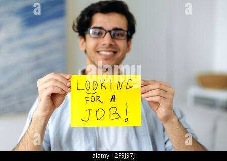 Défocused agréable et élégant homme de course mixte avec des lunettes, étudiant ou sans emploi, tenant un signe avec l'inscription à la recherche d'un travail, regardant la caméra, souriant amical Banque D'Images