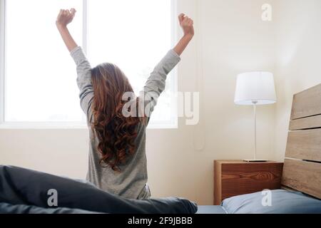 Yuong femme avec de longs cheveux bouclés assis dans le lit et s'étirant dans les premiers rayons du soleil Banque D'Images