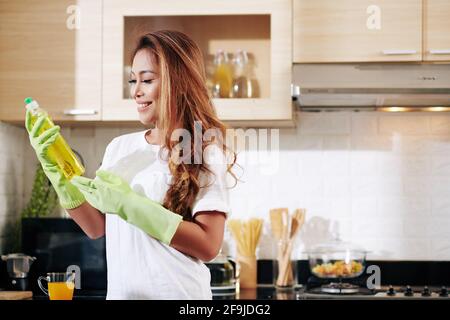 Souriante jeune femme vietnamienne dans des gants en caoutchouc regardant la bouteille sur le détergent de nettoyage ou le savon à vaisselle dans les mains Banque D'Images