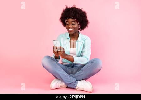 Femme africaine utilisant téléphone SMS assis sur le sol, fond rose Banque D'Images