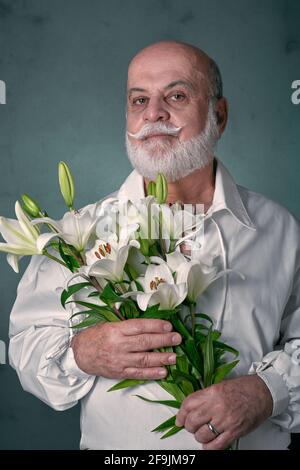 Portrait d'un homme âgé, chauve, avec une barbe grise et d'apparence latine, portant une chemise blanche et tenant un bouquet de lys blancs Banque D'Images