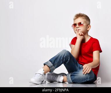 Le chef d'équipe blond élégant pour enfant est un t-shirt rouge, un Jean bleu, des baskets blanches et des lunettes de soleil Banque D'Images