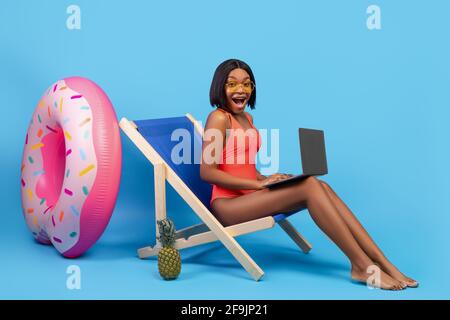 Soldes d'été. Femme noire excitée en maillot de bain assise dans une chaise longue avec ordinateur portable, criant OMG sur fond bleu Banque D'Images