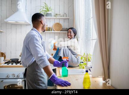 Jeune couple prenant la pause pendant le nettoyage de la maison, homme noir debout avec le détergent, petite amie assise sur la table de cuisine Banque D'Images