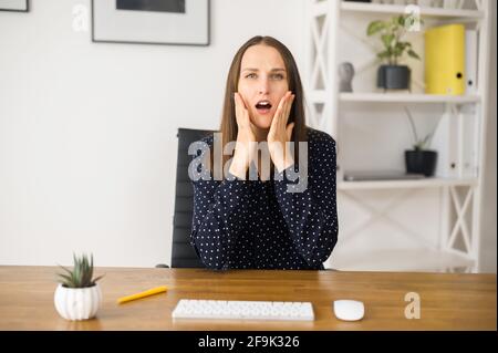 Une femme frustrée s'assoit au bureau et regardant la caméra avec une expression de visage choquée, malheureuse avec de mauvaises nouvelles la femme tient les joues avec les deux mains, a un problème avec le travail, la vue de webcam Banque D'Images