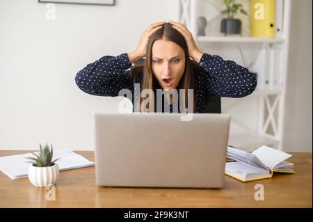 Une femme frustrée se trouve devant l'ordinateur portable et regardant l'écran avec une expression de visage choquée, mécontent de mauvaises nouvelles, la femme tient la tête avec les deux mains, a un problème avec le travail Banque D'Images