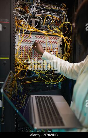 Gros plan vertical de la femme ingénieur réseau connectant les câbles dans l'armoire de serveur tout en travaillant avec le superordinateur dans le centre de données, espace de copie Banque D'Images