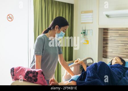 Fille dans un masque médical rendant visite à sa mère couchée au lit à l'hôpital. Banque D'Images