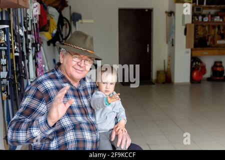 Portrait de personne homme âgé caucasien heureux senior assis à la maison de travail de garage avec cite petit enfant et en agitant la main bonjour geste. Grand-père et Banque D'Images
