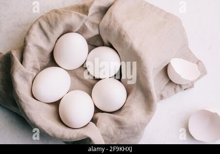 Panier d'œufs blancs sur un linge gris avec deux coques à côté sur fond de marbre blanc illuminées par la lumière de la vitre Banque D'Images