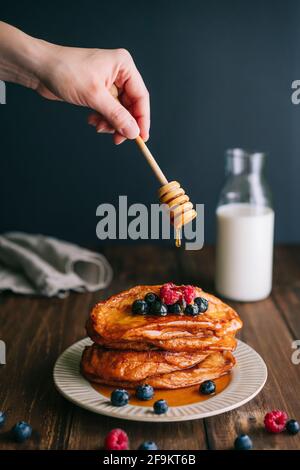 Faire à la main avec une cuillère à miel verser le miel sur des tranches de pain frites avec des myrtilles et des framboises dans une ancienne assiette beige sur une table en bois. Torrijas espagnoles Banque D'Images