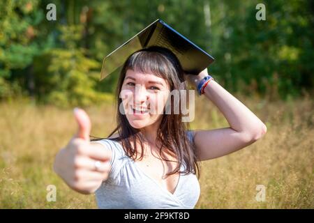 concept d'éducation en ligne. jeune femme heureuse élève tient un ordinateur portable au-dessus de sa tête comme une casquette universitaire carrée et montre un pouce vers le haut Banque D'Images