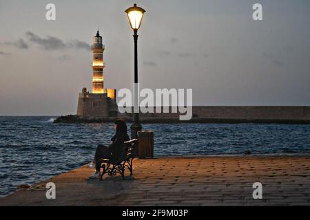 Paysage avec une fille assise sur un banc donnant sur le vieux phare au port vénitien de Chania en Crète, Grèce. Banque D'Images