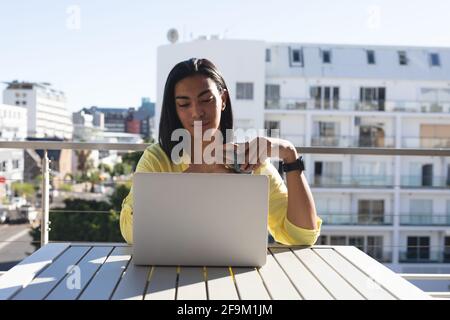 Femme souriante de race mixte transgenre assis à table sous le soleil terrasse avec ordinateur portable et café Banque D'Images