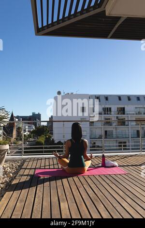 Vue arrière de la femme transgenre de race mixte pratiquant la méditation de yoga sur la terrasse du toit au soleil Banque D'Images