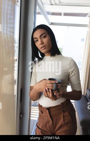 Portrait de la femme transgenre de race mixte debout par fenêtre tenue tasse de café Banque D'Images