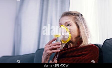 Une vieille femme vulnérable en quarantaine utilisant un inhalateur à oxygène et changeant de chanel à la télévision. Photo de haute qualité Banque D'Images