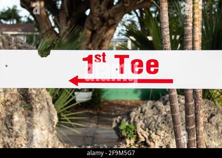 Un petit signe rectangulaire blanc avec des lettres majuscules rouges en gras qui orthographier 1st Tee. Flèche rouge pointant vers la gauche, l'arrière-plan est une jungle riche. Banque D'Images