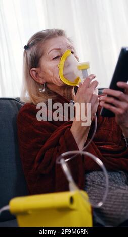 Une vieille femme vulnérable en quarantaine utilisant un inhalateur à oxygène et changeant de chanel à la télévision. Photo de haute qualité Banque D'Images