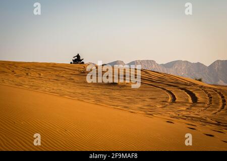 Silhouette d'un quad qui monte sur la dune de sable dans le désert, avec des traces de pneus derrière et des montagnes en arrière-plan, coucher de soleil, Fossil Rock, Sharjah, un Banque D'Images