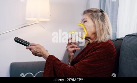 Une vieille femme vulnérable en quarantaine utilisant un inhalateur à oxygène et changeant de chanel à la télévision. Photo de haute qualité Banque D'Images