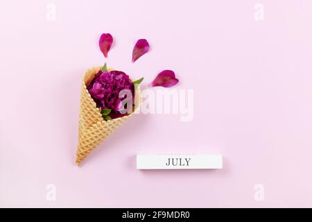 Fleur de pivoine en cône gaufré, pétales, bloc de bois avec inscription juillet sur fond de lilas pastel, espace de copie. Pose à plat minimaliste. Pour saluer c Banque D'Images