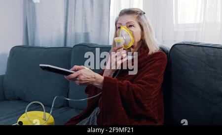 Une vieille femme vulnérable en quarantaine utilisant un inhalateur à oxygène et changeant de chanel à la télévision. Photo de haute qualité Banque D'Images