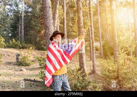 Jeune homme patriotique en chapeau de cow-boy tenant les Etats-Unis américain Drapeau et lever le poing et le bras dans la forêt.magnifique paysage de bois Au coucher du soleil.Indepe Banque D'Images