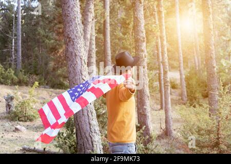 Homme en chapeau de cow-boy noir agitant le drapeau américain dans la forêt Au coucher du soleil.magnifique paysage boisé avec rayons du soleil et copier l'espace.United États indépendance d Banque D'Images