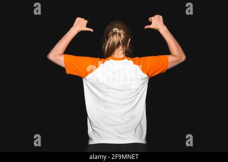 Une fille dans un T-shirt sur un fond noir pointe avec ses mains vers le dos, un endroit pour le texte. Concevez des vêtements pour espaces de copie. Banque D'Images