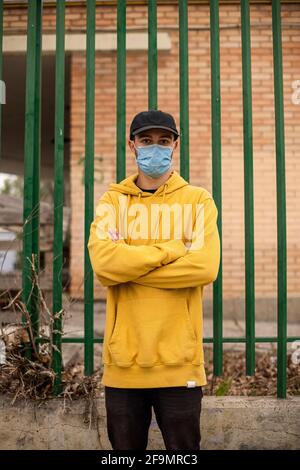 Jeune homme dans un pull à capuche jaune et un masque médical debout dans la rue avec les mains croisées Banque D'Images