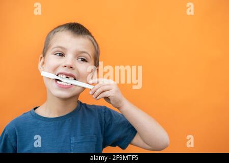 Garçon sans lait dent supérieure dans le t-shirt bleu tient brosse à dents dans sa bouche sur le fond orange. Banque D'Images
