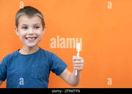 Un garçon sans la dent supérieure du lait dans un t-shirt bleu sourit et tient la brosse à dents à la main sur fond orange. Banque D'Images