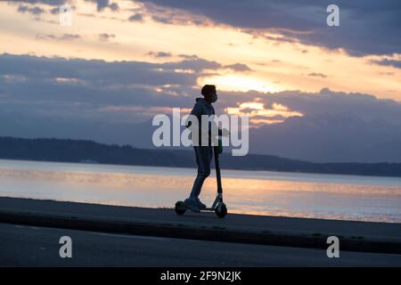 Seattle, Washington, États-Unis. 19 avril 2021. Un jeune homme portant un masque facial fait un scooter le long de la piste Alki tandis que le soleil se couche sur les montagnes olympiques de Seattle. Crédit : Paul Christian Gordon/Alay Live News Banque D'Images