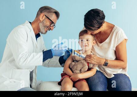 Sourire un médecin mature qui donne l'injection du vaccin à un garçon. Garçon se faire vacciner tout en étant assis avec sa mère. Banque D'Images