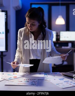 Manager stressé femme travaillant avec des graphiques de vérification de documents financiers, tenant des papiers tard dans la nuit. Femme d'affaires debout dans le bureau de démarrage faisant des heures supplémentaires pour respecter la date limite. Banque D'Images