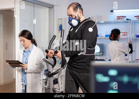 Athlète de performance en course sur cross trainer avec des électrodes fixées sur le corps et le masque dans le laboratoire d'essai, le médecin scientifique surveillant la fréquence cardiaque regardant la tablette pc. Banque D'Images