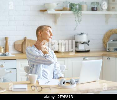 Besoin de repos. Femme mature fatiguée massant le cou tout en travaillant ou étudiant sur un ordinateur portable en ligne à la maison, assis dans la cuisine moderne à la maison. Concept de technologies et de personnes âgées Banque D'Images