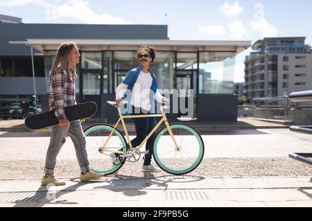 Deux amis de course mixte heureux de sexe masculin à vélo, portant un skateboard dans la rue et parlant. Mode de vie urbain vert, à l'extérieur comme à l'extérieur de la ville. Banque D'Images