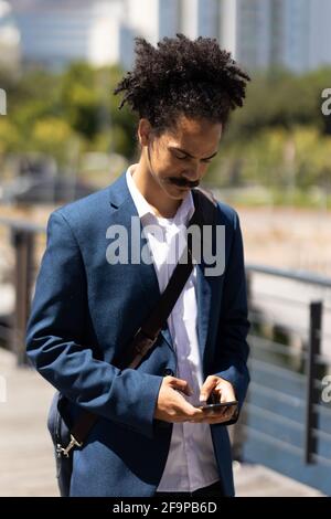 Homme de course mixte avec moustache, élégant, marchant dans la rue à l'aide d'un smartphone. Nomade numérique, en ville. Banque D'Images