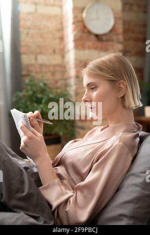 Jeune femme blonde allongée sur le lit et prenant des notes dans son ordinateur portable dans la chambre Banque D'Images