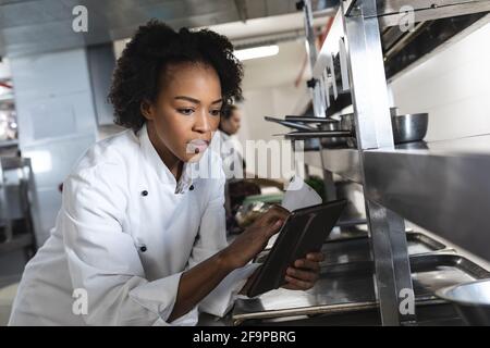 Chef professionnel de race mixte regardant une tablette avec un collègue en arrière-plan. Travaillant dans une cuisine de restaurant très fréquentée. Banque D'Images