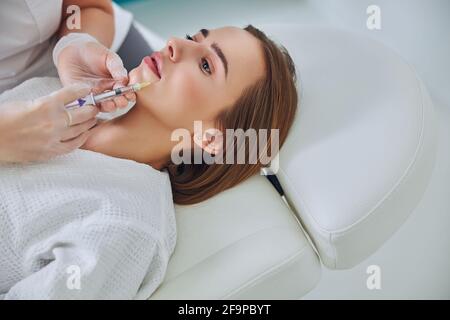 Charmante jolie dame couchée sur une chaise de médecine tout en recevant le sourire soulèvement et augmentation des lèvres dans le salon de spa Banque D'Images