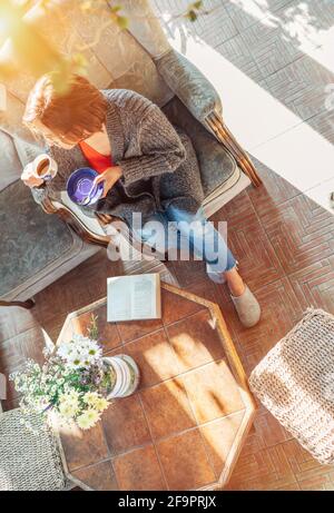 Vue de dessus d'une femme vêtue de vêtements confortables asseyez-vous dans un fauteuil confortable et buvez du café le matin maison véranda salon pendant qu'elle Banque D'Images