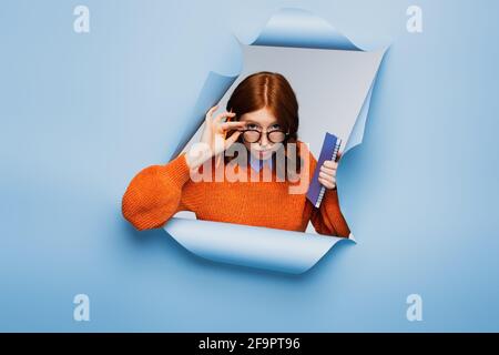 redhead jeune étudiant en chandail orange et lunettes tenant le carnet et crayon sur fond bleu déchiré Banque D'Images