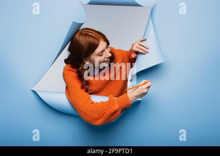 jeune femme joyeuse en chandail orange tenant un crayon près du bleu arrière-plan déchiré Banque D'Images